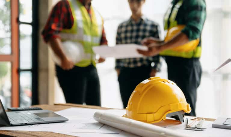 Construction worksite; close-up of a hard hat on a table while three engineers have a meeting in the background. Construction worksite; close-up of a hard hat on a table while three engineers have a meeting in the background.