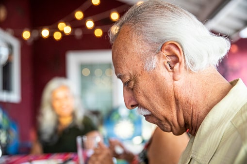 Man in a restaurant with his family, wearing a hearing aid and looking down