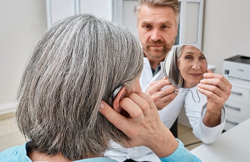 Senior woman trying on a hearing aid while looking into a mirror held by a hearing aid specialist in audiology clinic. Stylish hearing device Senior woman trying on a hearing aid while looking into a mirror held by a hearing aid specialist in audiology clinic. Stylish hearing device