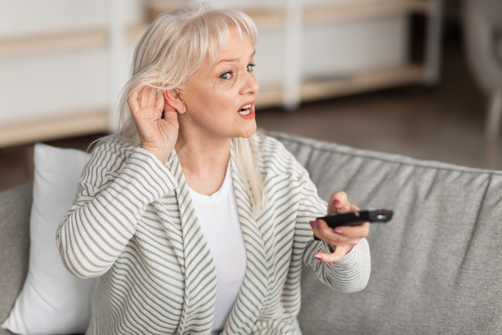 Portrait of \senior woman with impaired hearing watching TV, trying to listen holding her hand near ear to hear better, sitting on couch in living room.